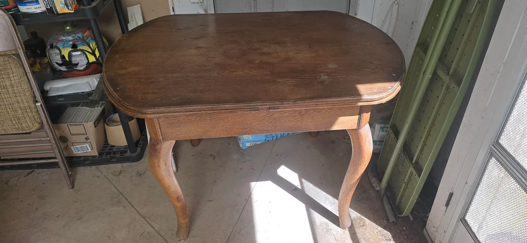 An old wooden table with curved legs sits in a sunlit room with concrete floors and various storage items on shelves nearby.