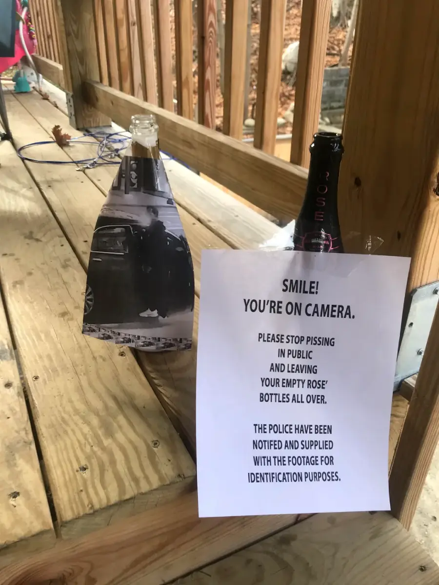 Two empty rosé bottles sit on a wooden porch. One has a black-and-white photo taped to it; the other has a note warning about security cameras and police involvement regarding public urination and bottle littering.