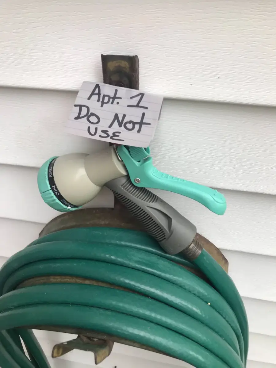A garden hose is coiled on a hanger mounted to a white wall. A nozzle is attached, and a handwritten sign above it reads, “Apt. 1 Do Not Use.”