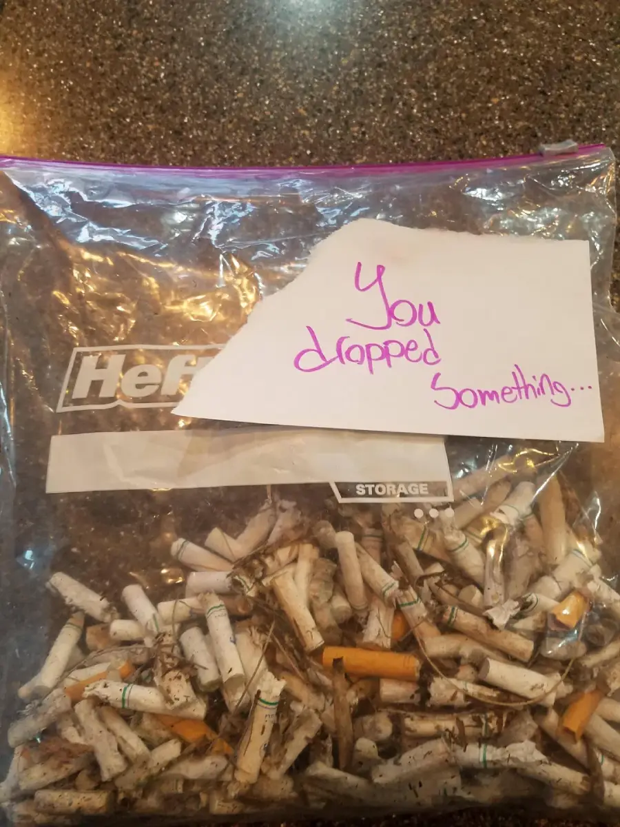 A plastic storage bag filled with used cigarette butts sits on a counter. On top of the bag is a piece of paper that reads, “You dropped something...” written in pink marker.