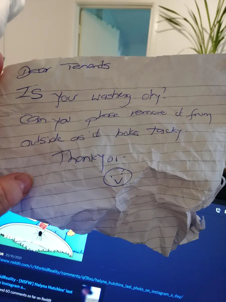 A handwritten note on crumpled paper asks tenants if their washing is dry and to remove it from outside as it looks tacky. A hand holds the paper, with a plant and computer screen in the background.