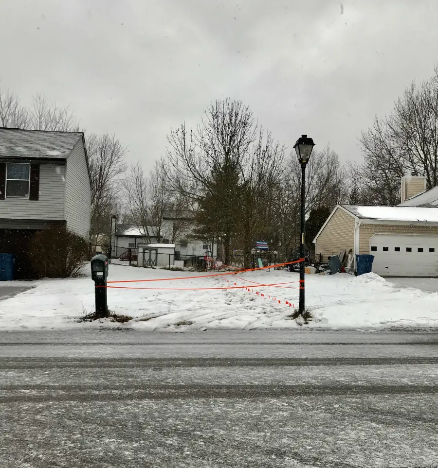 A snowy yard with orange string tied between a mailbox and a lamppost, creating a barrier across the walkway. Houses and bare trees are in the background under a cloudy sky.