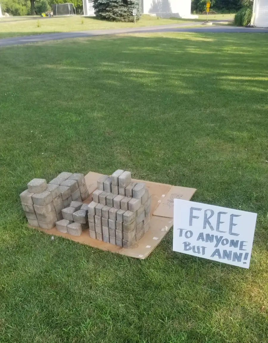 Stacks of rectangular concrete bricks on a lawn with a sign that reads, “FREE TO ANYONE BUT ANN!” The bricks are arranged on cardboard near a street, and houses are visible in the background.