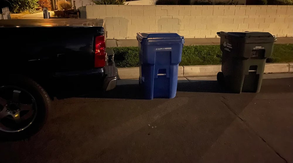A black truck is parked on a street next to a blue recycling bin and a green trash bin, which are placed at the curb at night.