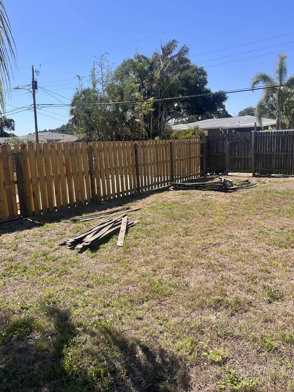 A backyard with patchy grass, a wooden fence, and a small pile of wooden planks on the ground. Trees and power lines are visible in the background under a clear blue sky.