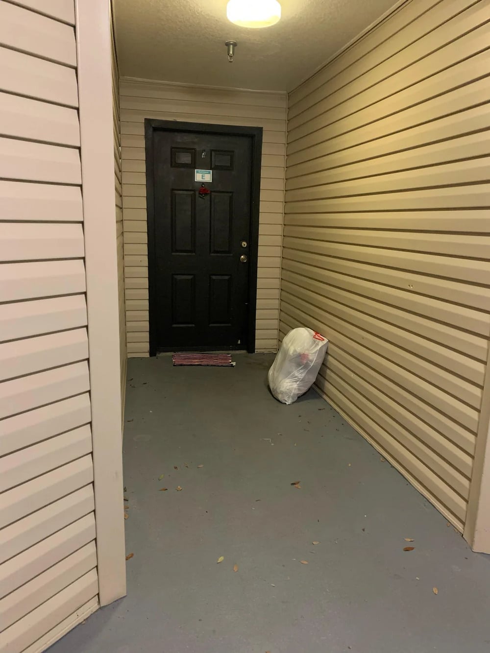 A black door at the end of a beige hallway with a ceiling light. A large white trash bag is placed on the floor near the door. Some leaves are scattered on the ground.