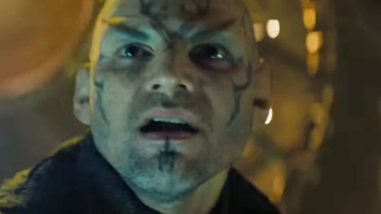 A man with intricate dark facial tattoos looks up with a shocked expression. The background is blurred with warm, yellow lighting.
