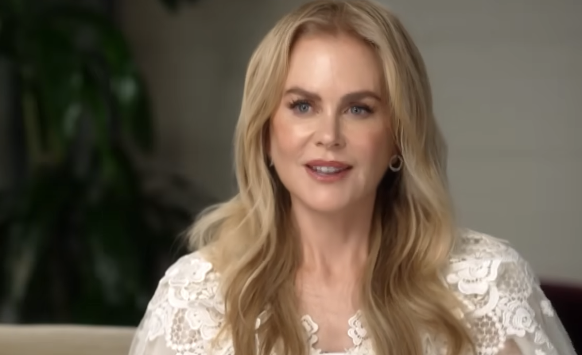 A woman with long blonde hair, wearing a white lace top, is sitting indoors and smiling. There is a blurred green plant in the background.