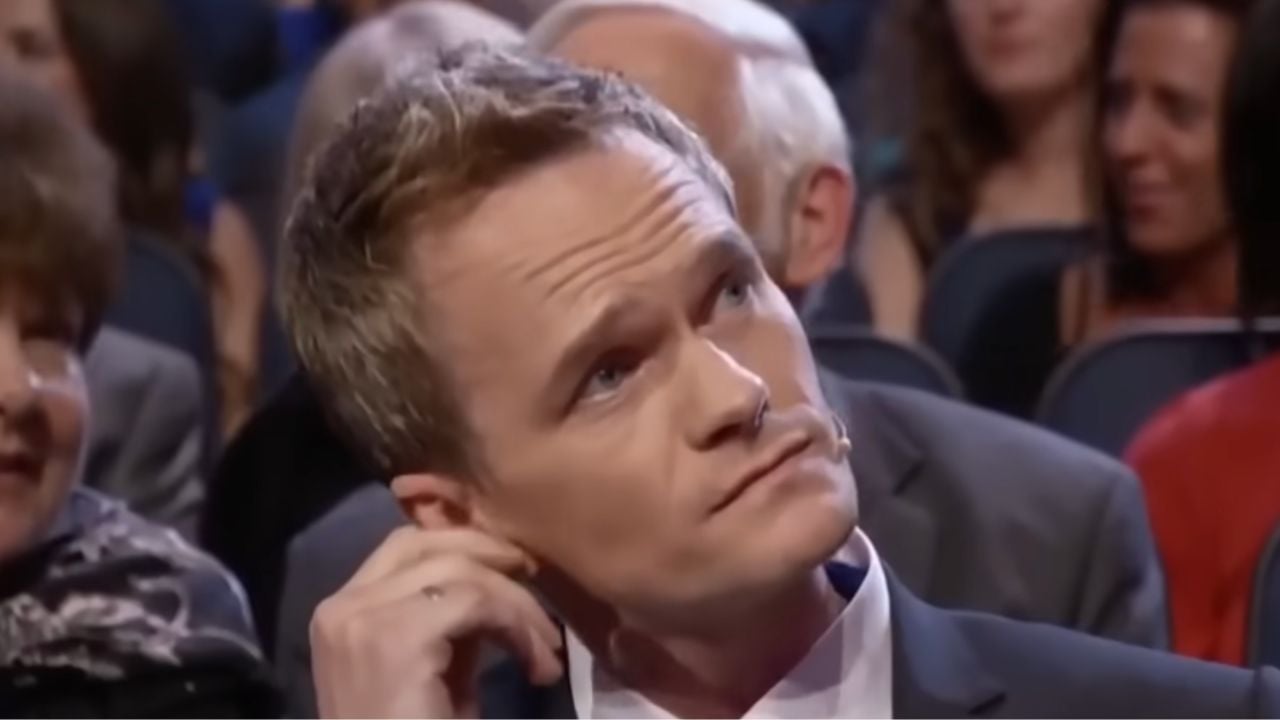 A man in a suit sits in an audience, looking slightly upward with a thoughtful expression while touching his ear. Other people are seated around him.
