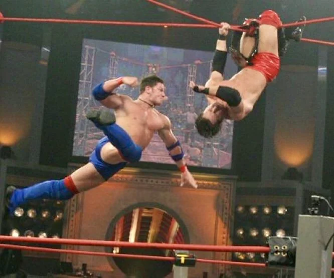 A wrestler in blue flies through the air delivering a kick to an opponent in red who is hanging upside down from the ropes in a wrestling ring.