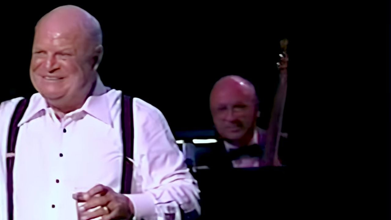 A smiling older man in a white shirt and suspenders holds a glass, while another man in a tuxedo sits behind him on a dimly lit stage.