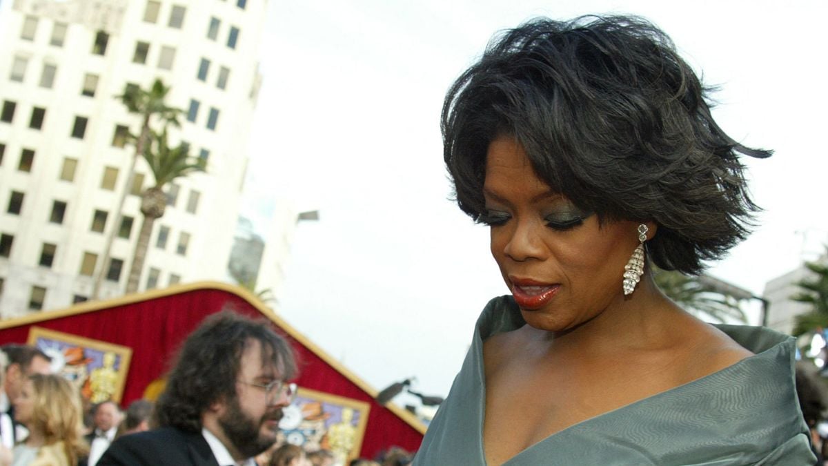 A woman in a green dress with elegant earrings looks down at an outdoor event, with people and a red tent in the background.