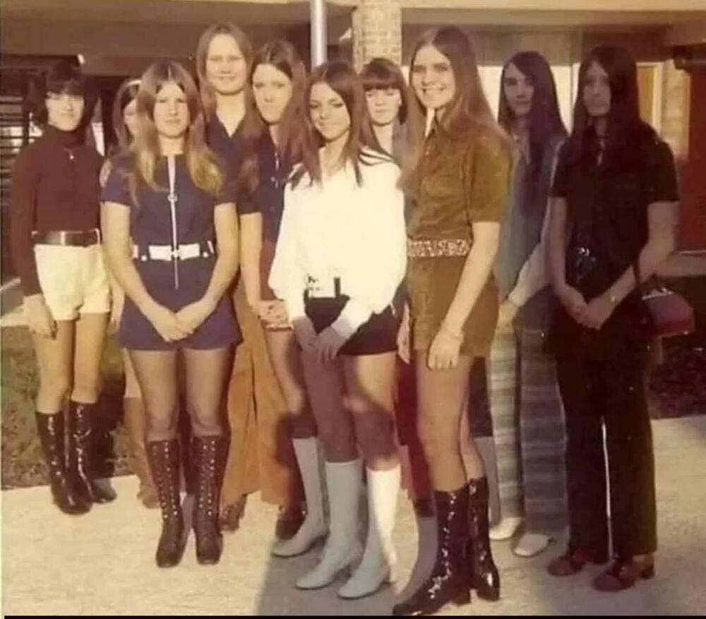 A group of nine young women pose outdoors in 1970s fashion, wearing mini skirts, dresses, and knee-high boots, with a building in the background.