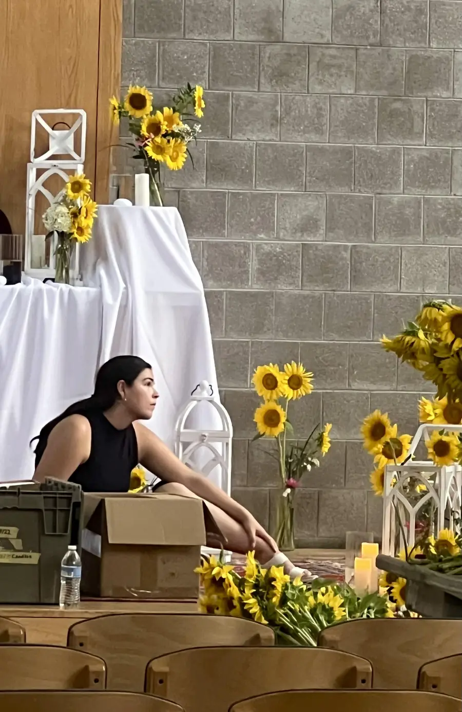 A woman sits on the floor surrounded by boxes, sunflowers in vases, and white lanterns. White cloth drapes over a table in the background against a gray brick wall.