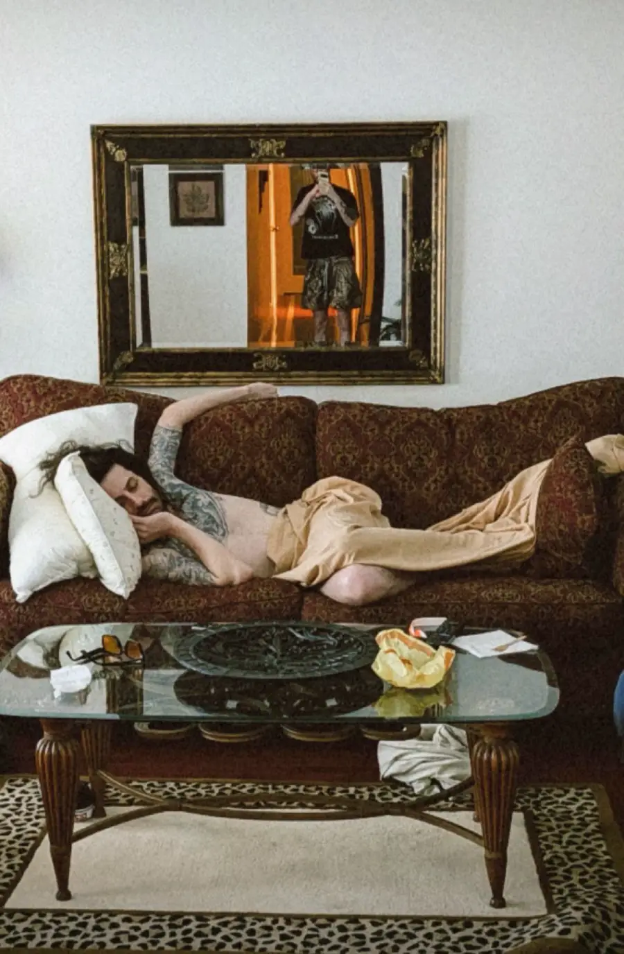 A person with tattoos sleeps on a couch with a blanket, while another person is seen photographing the scene in a large mirror hanging on the wall behind them. A glass coffee table and rug are in front.