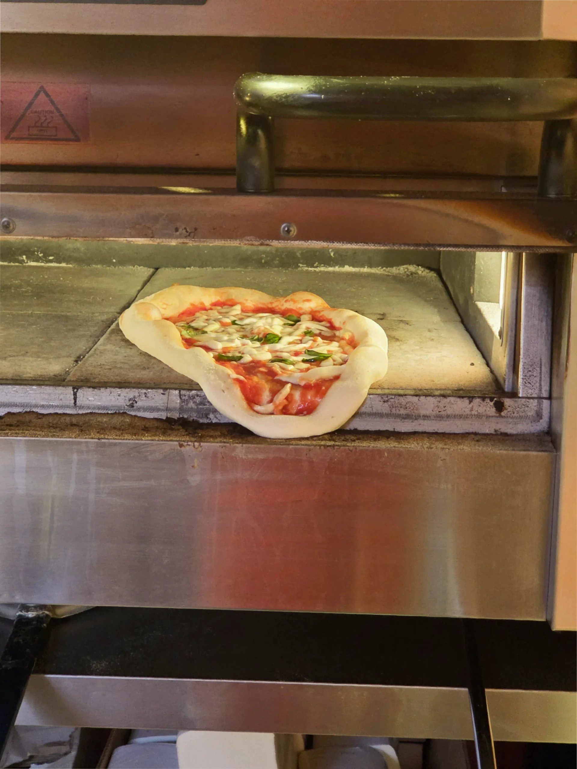 A pizza in an oven has sagged over the edge of the baking stone, with part of the dough hanging down, and cheese and toppings visible on top.