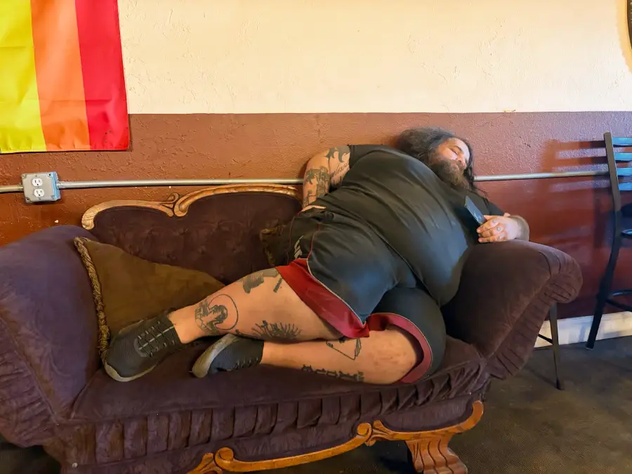 A person with tattoos and a beard lies on a dark purple couch, resting against a pillow. A rainbow flag hangs on the wall nearby, and chairs are visible in the background.