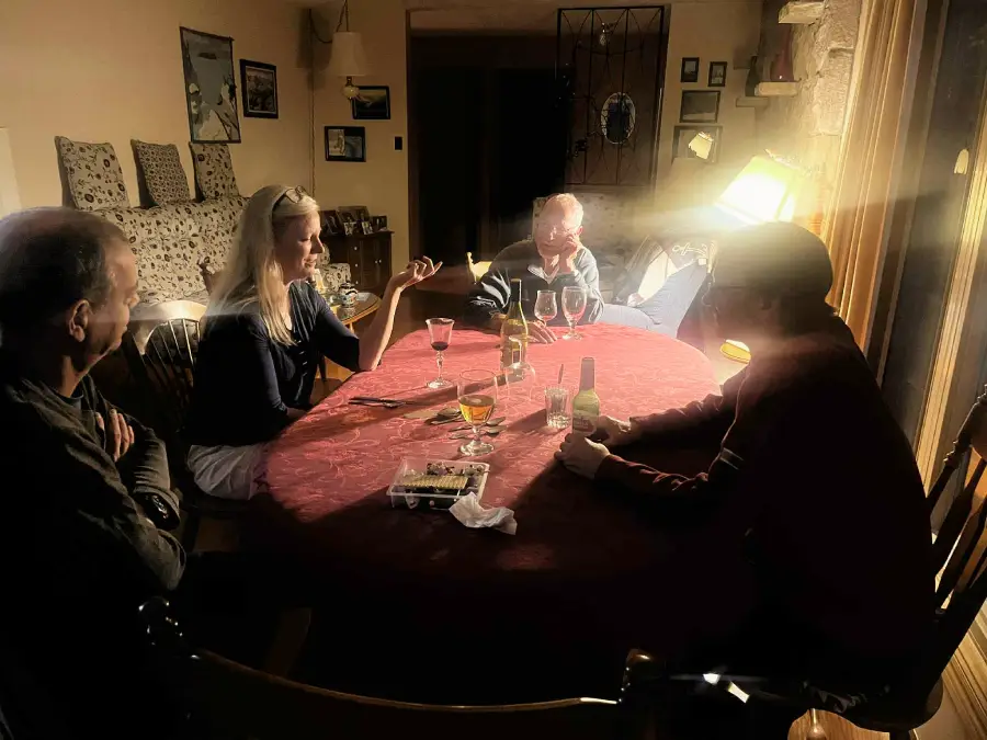 Four people sit around a round table with drinks and snacks in a dimly lit room. A bright lamp on the right casts dramatic light across the scene.