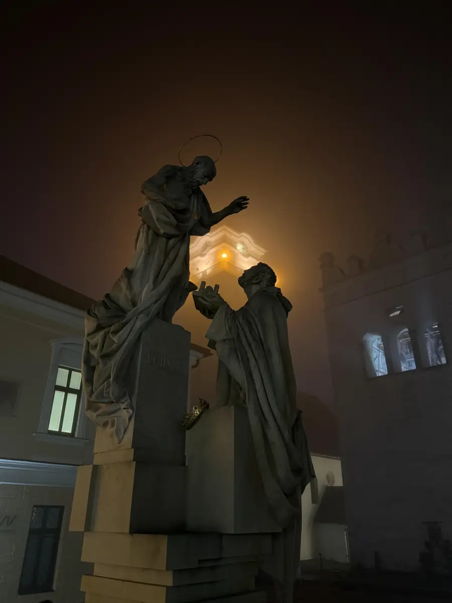 Two stone statues stand facing each other at night, illuminated by a warm streetlight behind them. Fog softens the light, creating a glowing halo effect, while surrounding buildings appear dimly in the background.
