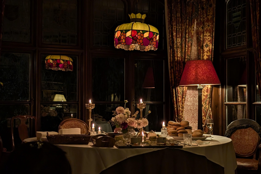 A warmly lit dining room with a table set for a meal, featuring candles, bread, flowers, and glassware. Stained glass and red lampshades provide cozy lighting in an elegant, vintage setting.