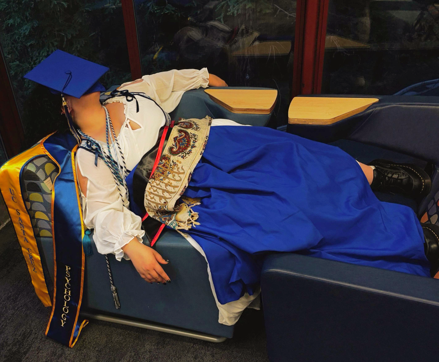 A person in graduation attire and a blue dress lies asleep on a chair, graduation cap covering their face. Sash reads “Psychology.” Small desks surround the chair; greenery is visible outside the window behind them.