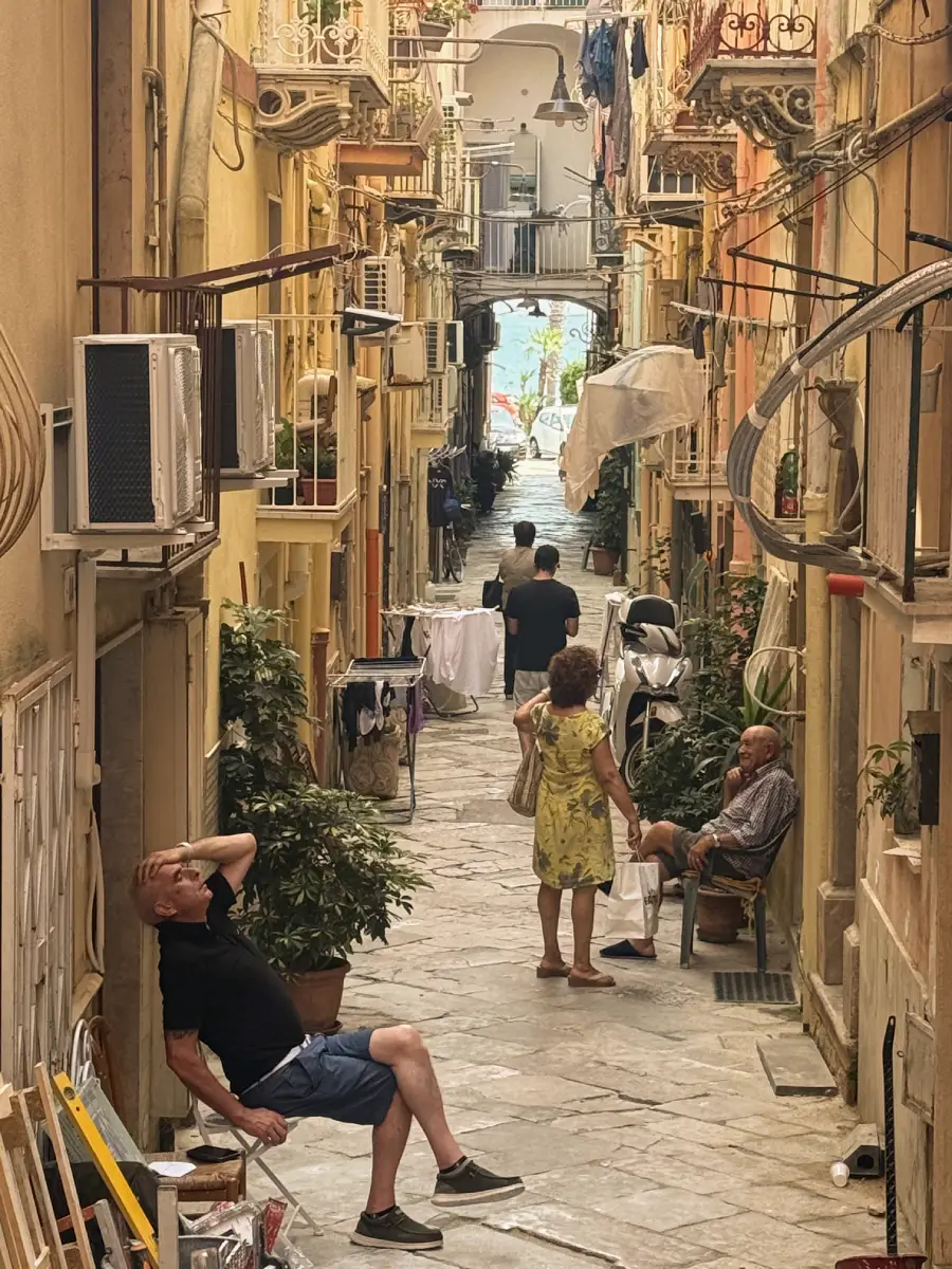 A narrow, sunlit alley with clotheslines, plants, and balconies. People walk or sit, casually relaxing. The scene feels lively and intimate, with colorful buildings lining both sides.