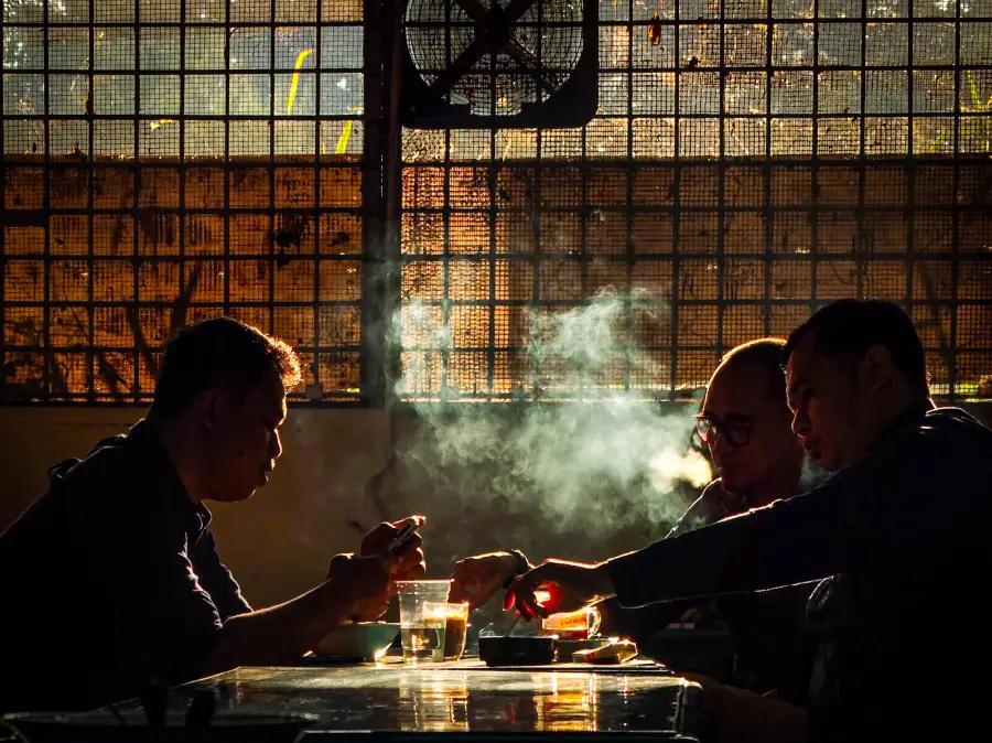 Three people sit at a table, eating and drinking with light streaming through a grid window. Smoke drifts in the air, creating a dramatic, moody atmosphere.