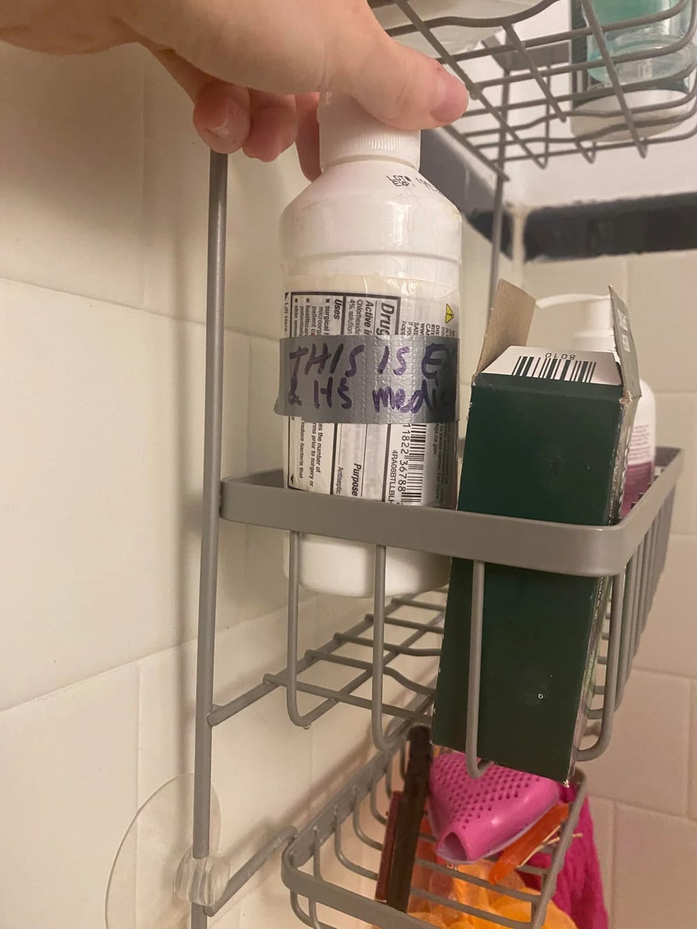A hand holds a white bottle labeled “THIS IS LH’S MEDICINE” on a shower shelf, next to a green box and pink items. White tiled wall in the background.