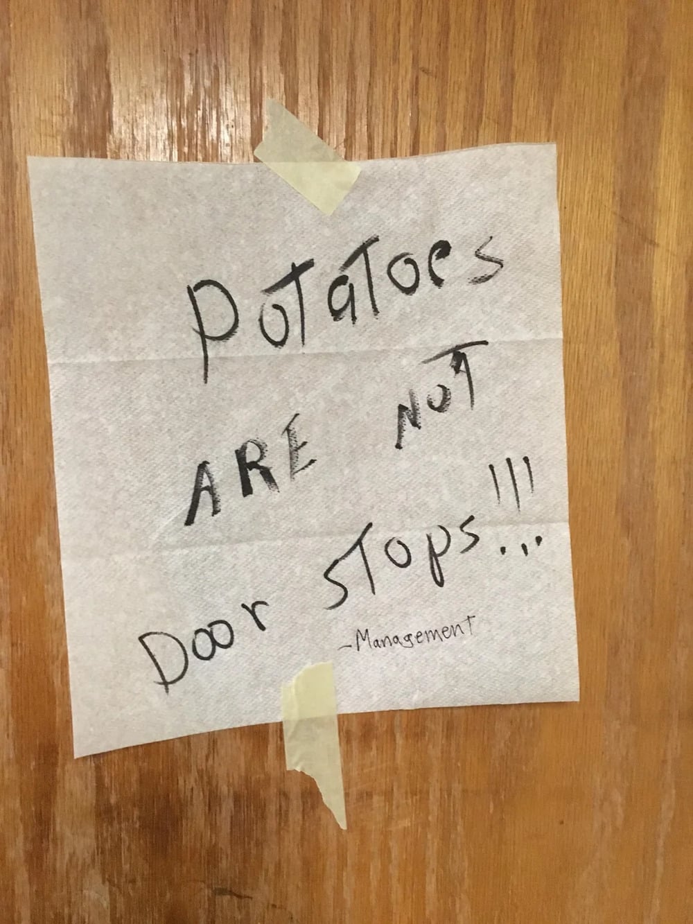 A handwritten sign on a wooden door reads: “Potatoes are not door stops!!! -Management.” The sign is taped at the top and bottom.