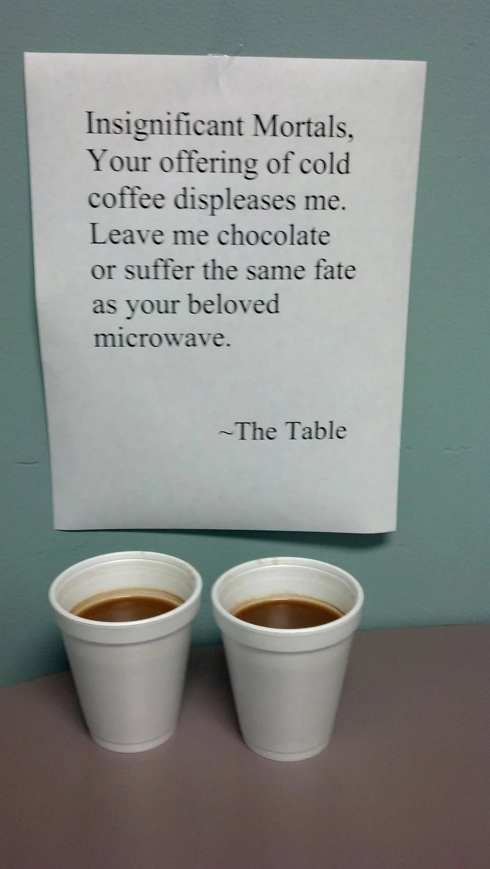 Two styrofoam cups of coffee sit on a table below a humorous printed sign requesting chocolate instead of cold coffee, signed “~The Table.”