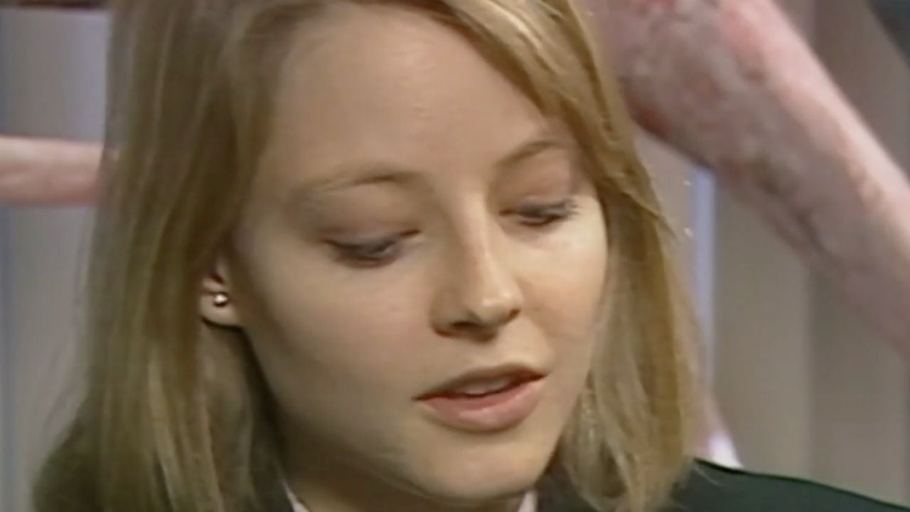 A woman with straight blonde hair looks downward with a neutral expression. She is wearing a dark jacket and small earrings. The background is blurred with light and pinkish elements visible.