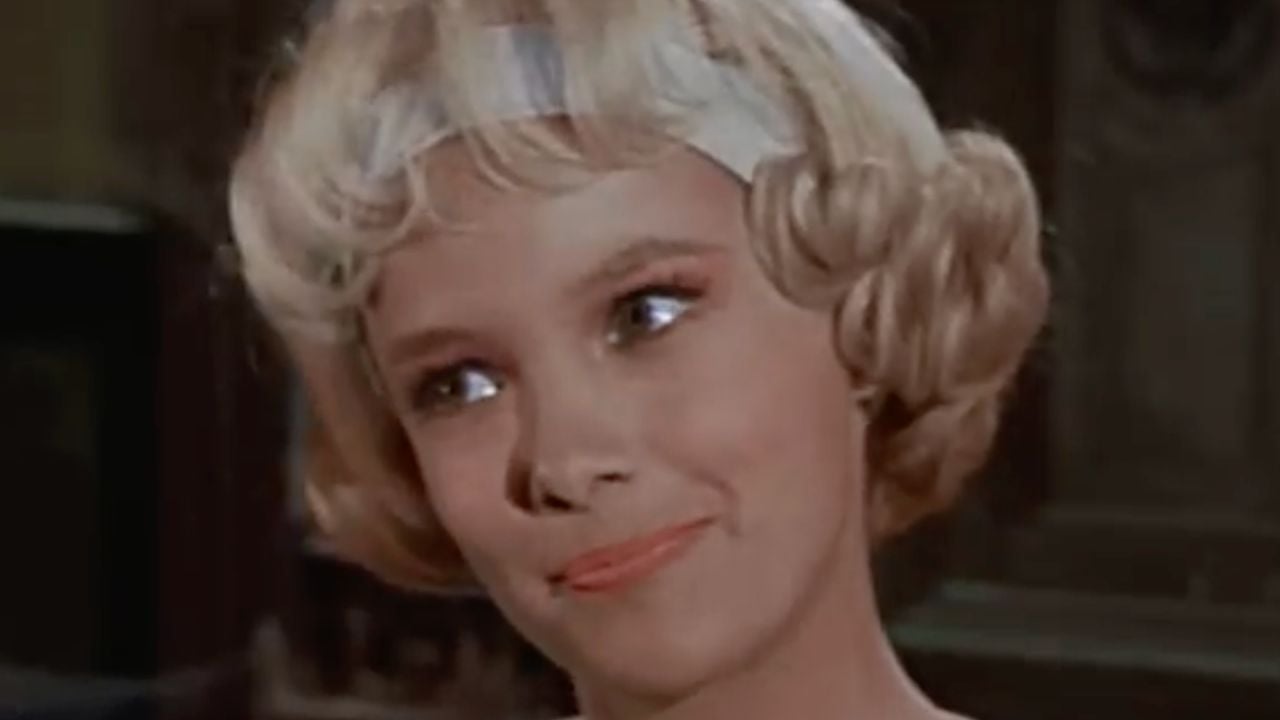 A woman with short, curly blonde hair and a white headband smiles softly, looking slightly to the side.