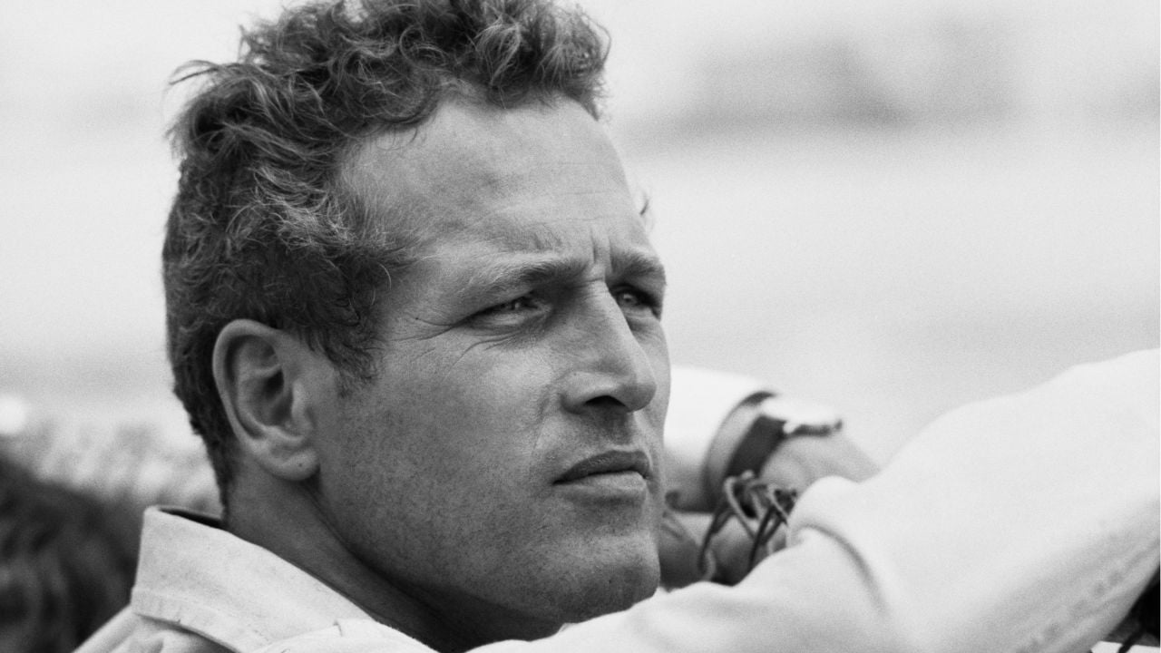 Black and white photo of a man with curly hair, looking thoughtful into the distance. He is wearing a light shirt, and his arms are resting on something in front of him.