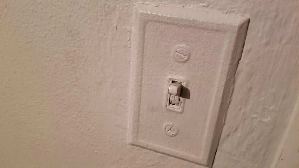A light switch and wall plate covered in white paint, blending into the similarly colored wall. The paint obscures some details of the switch and plate.