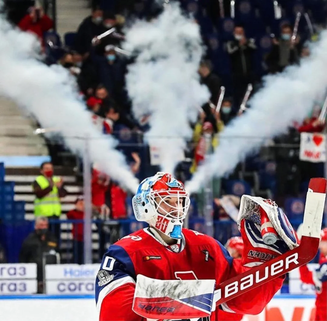 A hockey goalie in red gear stands on the ice as smoke cannons behind him create the illusion that smoke is coming out of his helmet. Fans are visible in the background, cheering and holding flags.