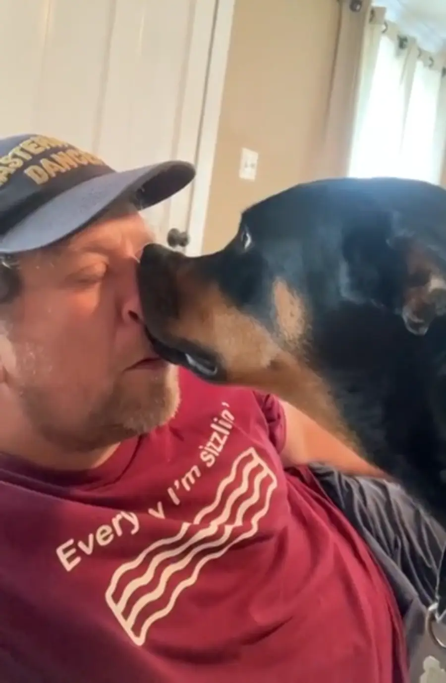 A man wearing a cap and a maroon shirt lounges while a black and brown dog affectionately touches his nose with its nose.