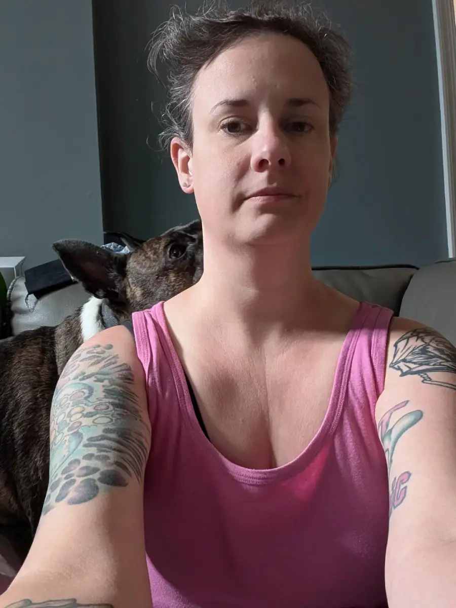 A woman in a pink tank top with tattoos on her arms sits indoors. A brown and black dog is behind her on the couch, partially hidden.