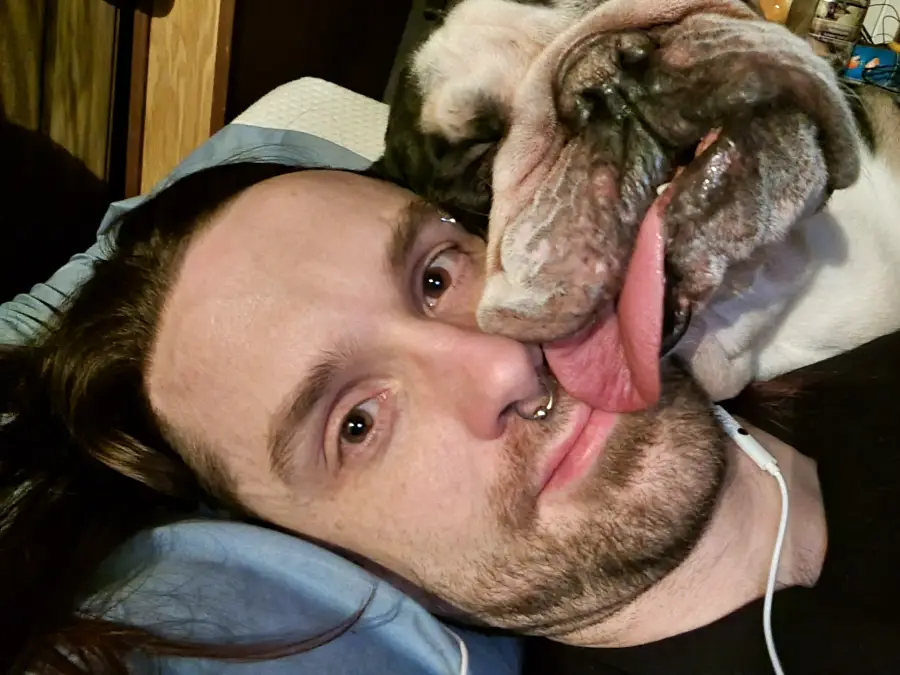 A man lies on a pillow while a bulldog rests its face on his, with its tongue hanging over his cheek.