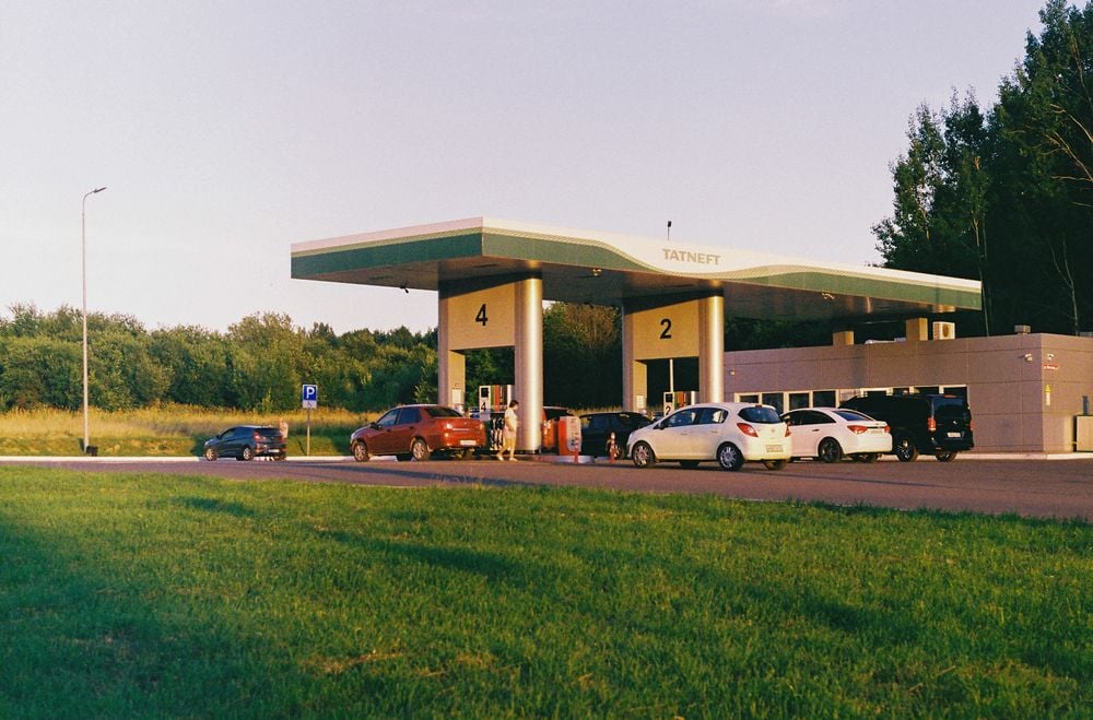 A gas station with multiple cars at the pumps, surrounded by grass and trees under clear skies. The station canopy displays “TATNEFT” and is labeled with pump numbers 2 and 4.