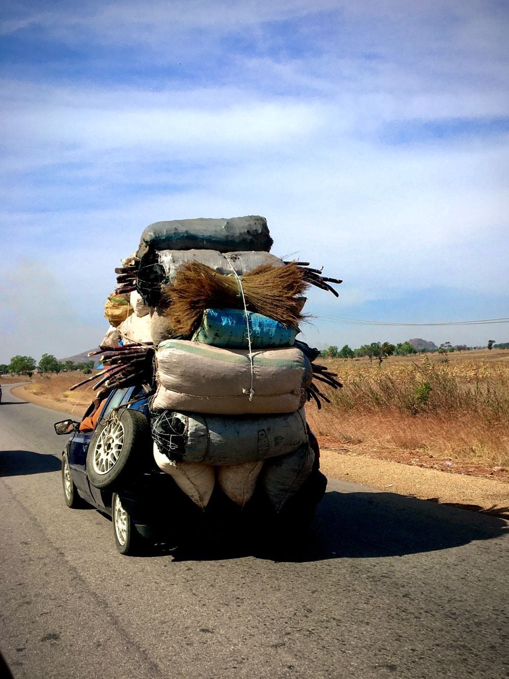 A small car is overloaded with mattresses, sacks, and bundles, some tied on top and sides. The car is driving on a rural road under a blue sky, with dry grass and trees in the background.