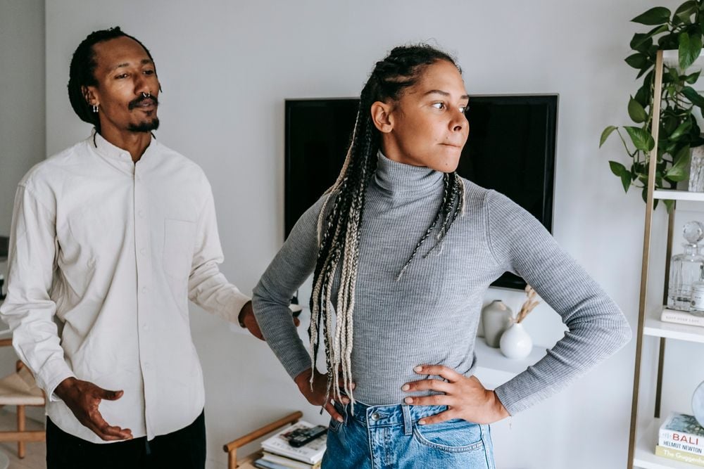 A man in a white shirt talks to a woman in a gray turtleneck who looks away with her arms on her hips, appearing upset, in a modern living room.