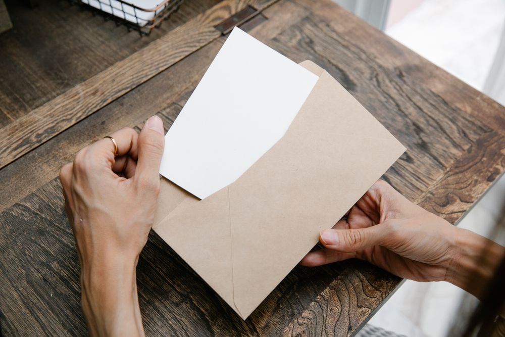 A person opens a brown envelope and pulls out a blank white card while sitting at a wooden table.