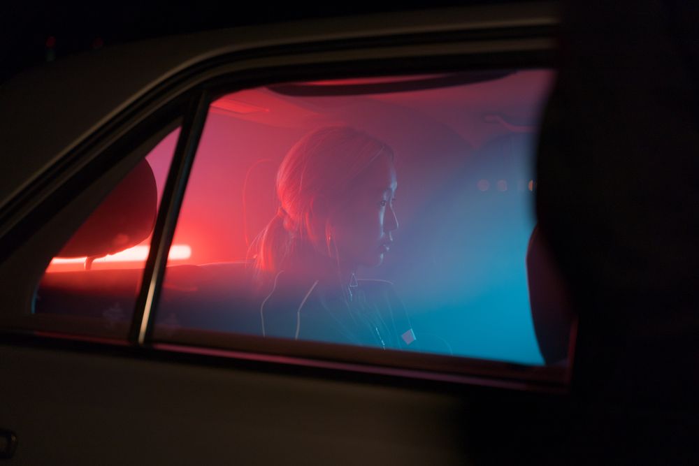 A person sits in the backseat of a car at night, illuminated by red and blue lights from outside, creating a moody atmosphere through the window.