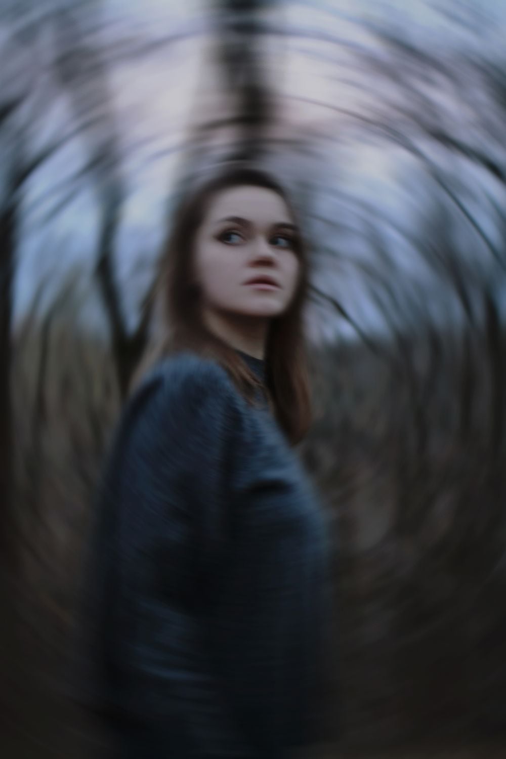 A woman stands outdoors in a dark sweater, surrounded by twisted, blurred trees, creating a swirling, dizzy effect in the image.