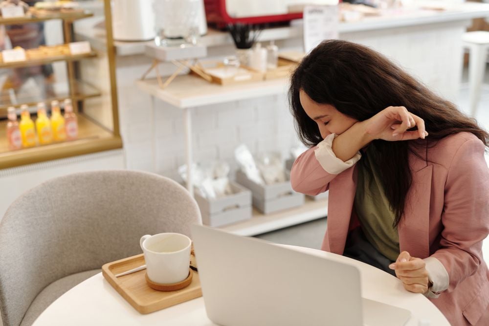 Woman sitting at a table with a laptop and a cup, covering her mouth with her elbow as she coughs or sneezes, in a bright café setting.