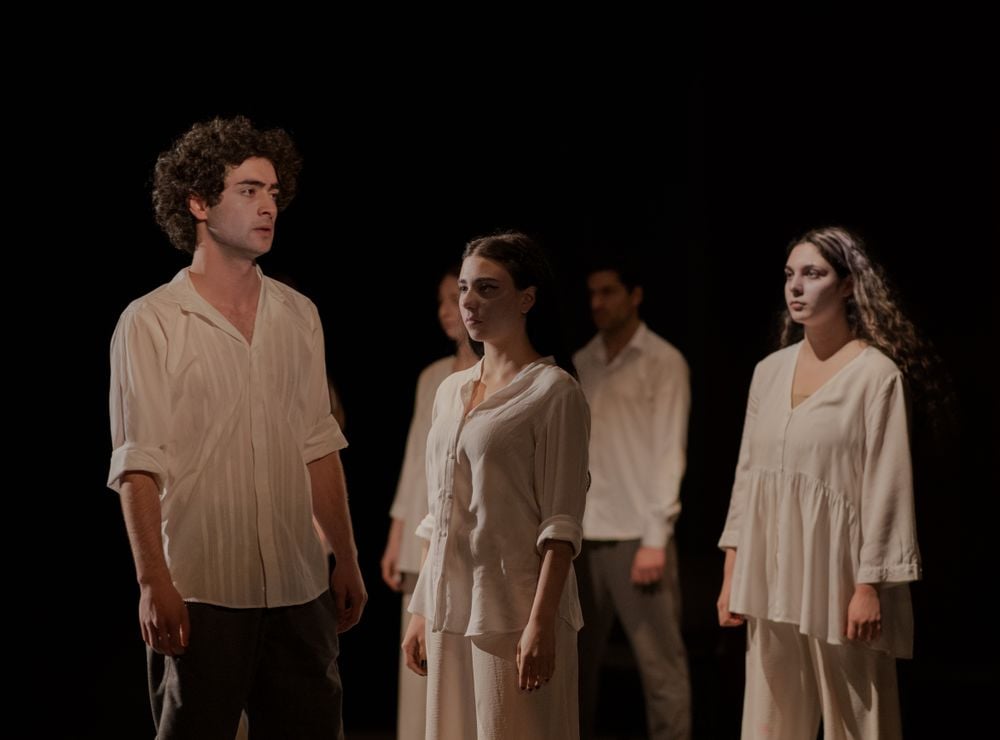Four people in loose, pale clothing stand on stage with serious expressions. The lighting is dim, highlighting their faces against a dark background. They appear to be performing a dramatic scene.