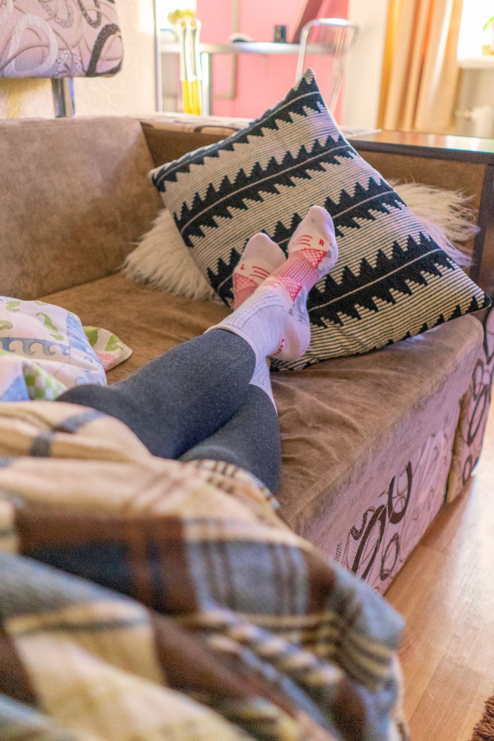 A person wearing cat-themed socks and leggings lounges on a couch with patterned pillows, covered partially by a plaid blanket in a cozy living room.
