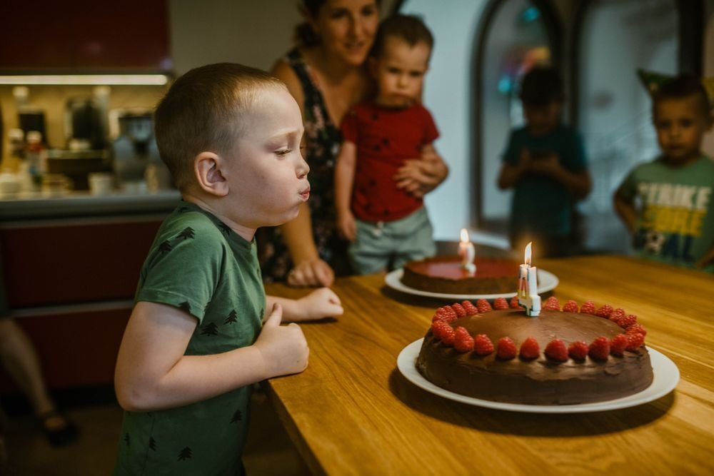 A young boy blows out candles on a chocolate cake with raspberries, while a woman and other children watch in a kitchen, celebrating a birthday.