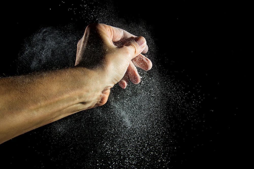 A hand snaps fingers against a black background, sending a burst of fine white powder or dust particles into the air.
