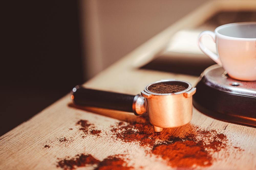 A portafilter filled with ground coffee rests on a wooden surface, surrounded by scattered coffee grounds. A white cup sits nearby on a saucer.